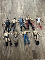 Photo showing nine WWF/WCW wrestler action figures arranged on a wood floor with visible wear and variances in condition including one missing ponytail and chew marks on a foot