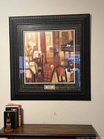 Framed art print showing a cafe or bar interior scene with chairs and tables, mounted on a white wall above a dark wooden table with books and a small box on it.