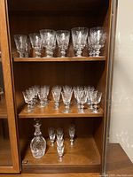 Full view of cabinet shelves holding two rows of stemmed crystal glasses on top and bottom shelves, clear with etched design.