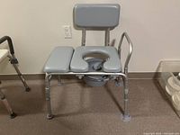 Grey padded commode with backrest, armrest, open center seat, and adjustable aluminum legs on indoor carpeted floor.