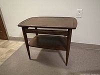 Photo of a wooden side table with a warm brown finish, showing top surface, tapered legs, and lower shelf against a plain wall and carpeted floor.