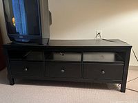 Black-brown IKEA Henmes TV stand with 3 drawers and 3 open shelves, sitting against a beige wall, with a TV placed on top.