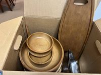 Open cardboard box containing several round wooden dishes and bowls stacked inside. Two wooden cutting boards including one with handle are visible standing upright. Metal pan with handles is also included in the box.