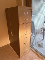 Beige metal filing cabinet with four drawers, label holders, and key lock. Standing on carpet by window.