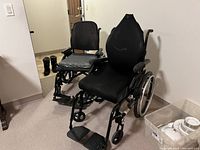Photo shows two manual wheelchairs side by side in a room corner. The closer wheelchair has padded seat and backrest, larger wheels and padded armrests. The second wheelchair behind has mesh seat and backrest and exhibits more wear.