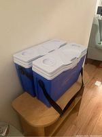 Pair of blue Rubbermaid slim coolers with white lids placed on wooden bench, showing black shoulder straps and cup holders on lids.