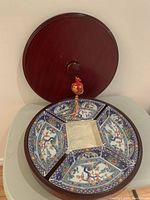 Top view showing varnished circular wooden case lid with a knob and attached red decorative tassel with gold details.