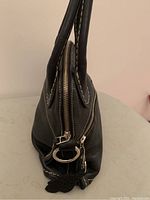 Side profile showing black leather purse with sturdy handles, white decorative stitching, zippered top closure, and attached leather fob.