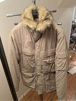 Front view of Guess beige/tan parka jacket with faux fur hood showing front pockets and zipper closure.