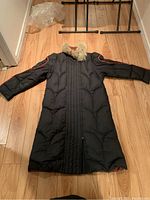 Front view of a full length black down parka laid flat on wood floor showing fur-lined hood and zipper.