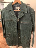 Front view of green suede Liz Baker jacket on a hanger, shows zippered chest and waist pockets and button closure