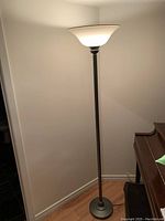 Full view of the torchiere floor lamp standing against a wall showing its base, pole, and glass shade illuminated.