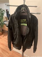 Black Super Dry JPN Moody Norse Bomber jacket hanging on a hanger showing front view and fleece inner lining.