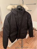 Front view of black Woods men's bomber jacket with parka hood and beige faux fur trim, showing front zipper and flap pockets.