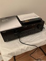 Yamaha HTR-6050 AV receiver amplifier with remote control and manual on top, showing front panel with control buttons, headphone jack, and volume knob. Power cord attached.