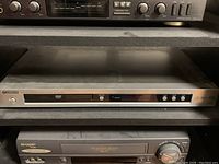 Silver Yamaha DVD player DV S5750 on a shelf below an audio receiver unit and above a Sharp cassette player.