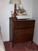 Photo of antique wooden chest of drawers with three drawers and round knobs, standing on turned legs, placed against a white wall on a carpeted floor.