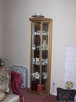 Photo of tall wooden curio cabinet with glass panels showing display of ceramic and porcelain figurines, cups, and small decorative items on several shelves.