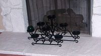 Photo showing black decorative metal candle stand with multiple candle holders placed in front of a fireplace.