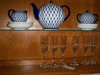 Photo showing china teapot, creamer, sugar bowl on saucers and plates with six clear stemmed glasses below on wooden shelf
