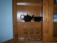 Photo showing a wooden cabinet shelf with a black china tea set (teapot and two tea cups) on the top shelf and three clear glass goblets arranged on the lower shelf.