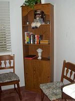 Wooden corner bookshelf in a kids' room with stuffed monkey, books, small candles, and decorative vase on shelves. Two chairs with floral cushions sit near the shelf.