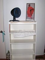 White wooden bookshelf with three shelves, a small desk fan and a framed picture on top