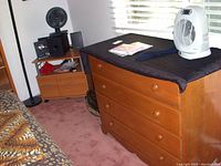 Photo showing a wooden dresser with three drawers, black protective cloth on top, various items placed on top, and a corner shelving unit nearby.