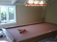 Photo of complete pool table set showing the table with pink felt, racked balls, cues on wall rack, and overhead lamp.