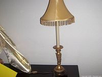 Photo shows a decorative table lamp with beige fabric shade trimmed with bead fringe, and an ornate gold-tone base with intricate detailing, set on a dark surface.