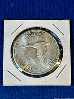 Close-up photo of reverse side of 1967 Canada Centennial silver dollar coin showing flying Canada goose with inscriptions "DOLLAR" and "CANADA 1867-1967." Coin is secured in a white 2x2 holder with staples.