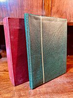 Photo showing two hardcover stamp albums, one green and one burgundy, standing upright on a wooden surface.
