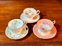 Three Aynsley English bone china teacups with matching saucers displayed on wood surface. Each set has distinct colors and patterns including pink gold floral, blue with clover, and pastel floral.
