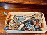 Box containing various car badges, hood ornaments, and parts arranged loosely on a wooden floor.