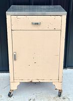 Front view of vintage metal medical cabinet showing drawer, door, caster wheels, and stainless steel top.