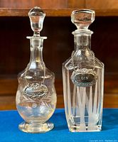 Both vintage decanters presented side by side on blue fabric, showing glass stoppered tops and metal label tags.