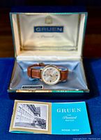 Vintage Gruen Precision men's wristwatch shown inside its original blue and white satin-lined presentation box with gold embossing. New old stock brown leather band attached. Accompanied by original Gruen informational booklet.