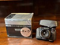 Photo of the Miranda dx-3 original box next to the camera with leather case set on a wooden surface. Box shows wear and the camera is inside the protective case.