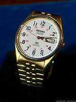 Close-up of the watch face showing the white dial with black and red numerals, day-date window, and gold tone casing and band.