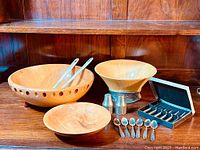 Wide shot of wooden bowls, tongs, salt and pepper shakers, and boxed set of cocktail forks on wooden surface.