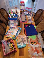 Overview of assorted arts and crafts supplies on a table including paint bottles, brushes, workbooks, colored paper, scissors, glue, markers, and craft jar.