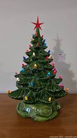Front view of green ceramic Christmas tree with multi-colored lights and red star on top on wooden surface against plain wall.