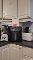 Three kitchen appliances on kitchen countertop: Oster blender, Cosori air fryer with manual, Sunbeam toaster.