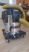Front and side view of DeWalt wet/dry vacuum with yellow hose attached, showing wheels and DeWalt logo on stainless steel tank.
