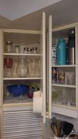 Two white kitchen cabinet doors open showing shelf with glassware, mixing bowls, jars, and travel mugs.