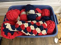 View of unfinished crocheted blanket in a plastic bin with multiple skeins of yarn in red, navy blue, and cream colors.