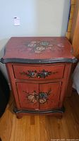 Front view of hand painted floral nightstand showing drawer and cabinet doors, painted deep red with black edges and floral decoration.