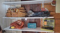 Photo showing the Mastercraft leather tool belt with hand tools, the black Mastercraft angle grinder with grinding and cutting discs, orange extension cord, Makita drill bit case, and Radley hole saw set on a white shelf.