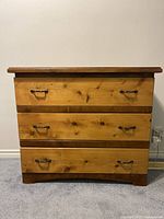 Front view of natural wood dresser with three drawers and metal handles, showing natural wood grain and minor wear.