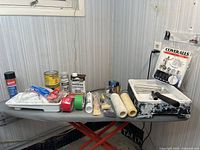 Photo showing various painting supplies on a table including paint rollers, paint trays, stir sticks, tape rolls, pigment cans, and disposable coveralls.